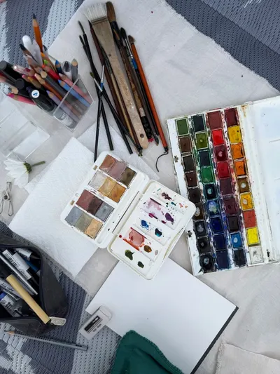 A top view of watercolor art supplies including palettes, brushes, pencils on a textured surface.