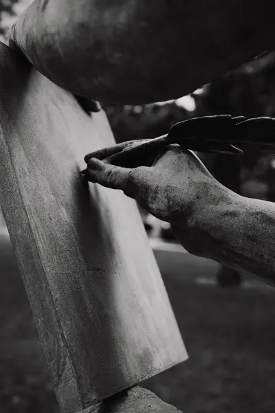 Black and white photo of a statue's hand holding a quill, writing on a tablet.