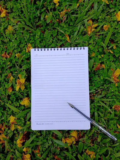 A blank lined notepad with a pen lies on vibrant green grass with yellow flowers.