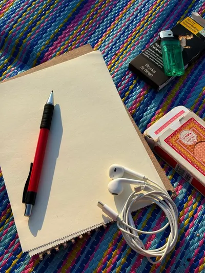 A colorful outdoor setting featuring a notebook, pen, earbuds, playing cards, and a lighter on a vibrant mat.