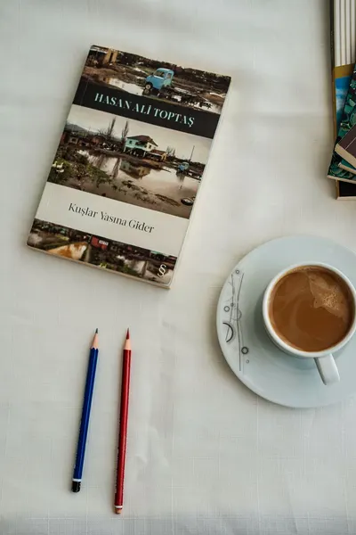 A book, coffee cup, and colored pencils on a white tablecloth create a cozy reading scene.
