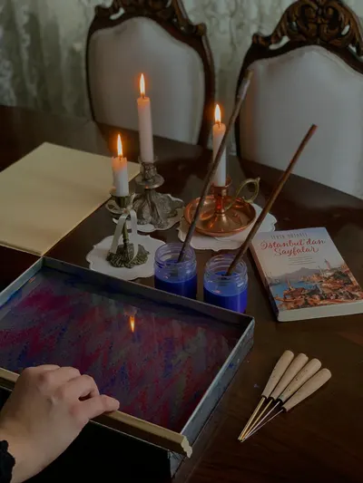 Ebru marbling art setup with vibrant colors, lit candles, and a book on a wooden table.