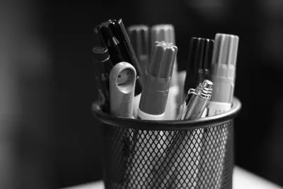 A monochrome close-up of pens and markers in a mesh holder on a desk.