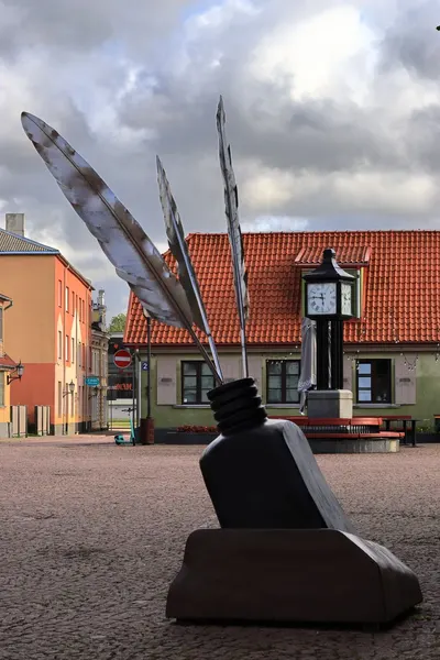 A town square features a large inkwell sculpture with feathers under a cloudy sky.