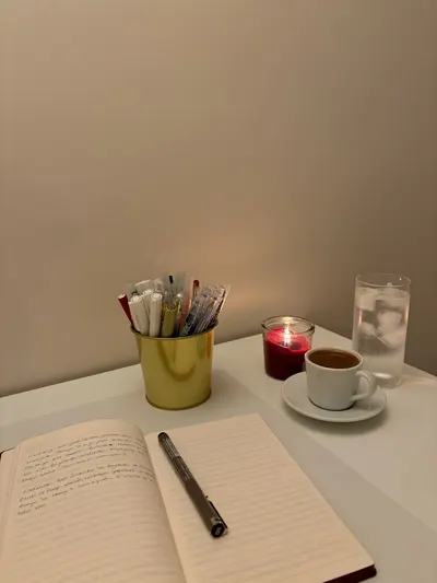 A calm workspace setup featuring a notebook, pen, coffee, candle, and glass of water at night.