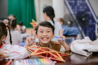 Smiling child holding peace sign with colorful pens and art project in classroom setting.