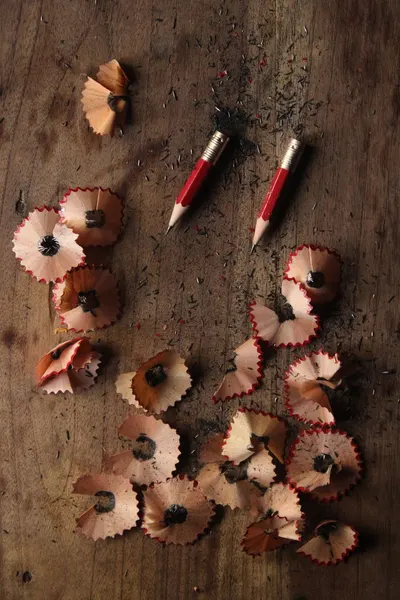 Two red pencils surrounded by wooden shavings on a rustic wooden table.