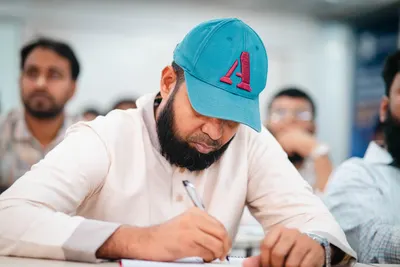 A man wearing a cap is intensely writing notes in a classroom, showing focus and education.