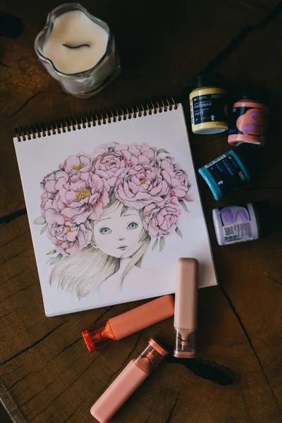 Notebook with illustrated girl adorned with pink flowers, surrounded by art supplies.
