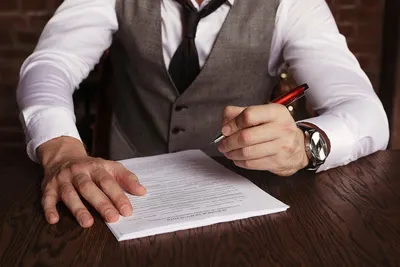 Business professional in vest signing important contract at a desk with red pen.