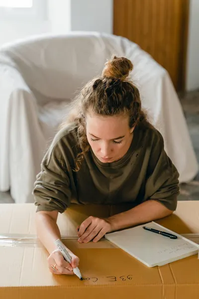 From above of concentrated young woman checking and taking notes on cardboard box while preparing for relocation