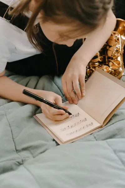 A young musician writes music notes in a notebook beside a saxophone, practicing creativity.