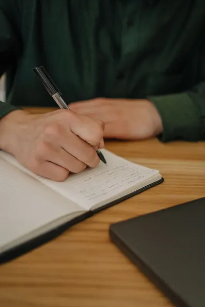 Close-up of a hand writing notes in a notebook on a wooden desk indoors.