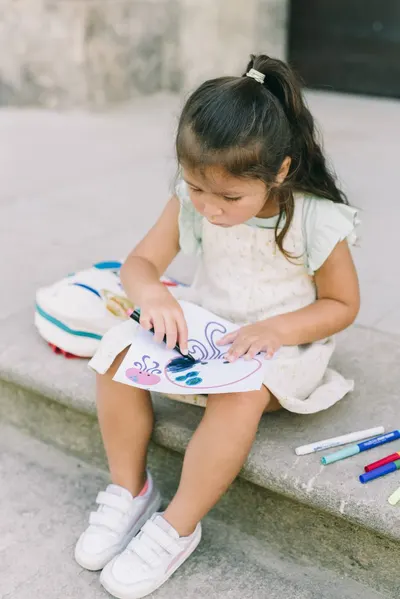 Adorable child coloring on paper with markers, seated outside on a concrete step.