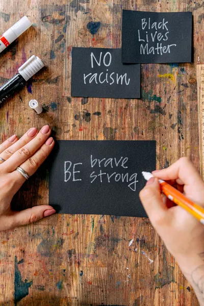 Handwriting encouraging messages like 'Be Brave' and 'No Racism' on black cards with focused hands.