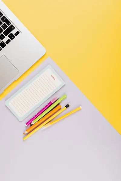 A flat lay of colorful stationery with a laptop on a vibrant yellow and gray desk.