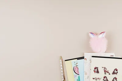 Flat lay of colorful notebooks, pen, and fuzzy pom-pom on a beige desk with ample copy space.