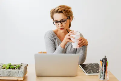 Focused woman working on laptop while holding coffee cup in office environment.