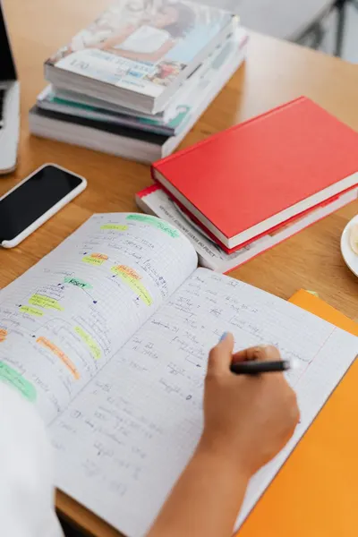 Student writing notes in a notebook during study session with books, phone, and highlighters.