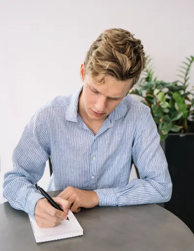 Focused young man writing notes on a notepad at a table indoors.
