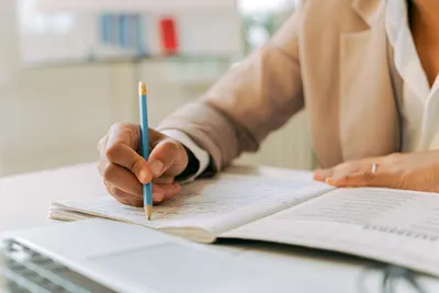 Close-up of a person in a beige blazer writing in a notebook in an office environment.
