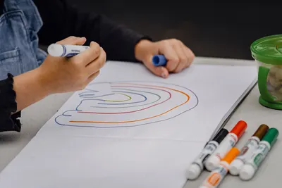 Crop unrecognizable kid holding marker while sitting at table and painting rainbow on sheet of paper