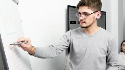 Confident young man with eyeglasses writing on a whiteboard during a business strategy presentation.