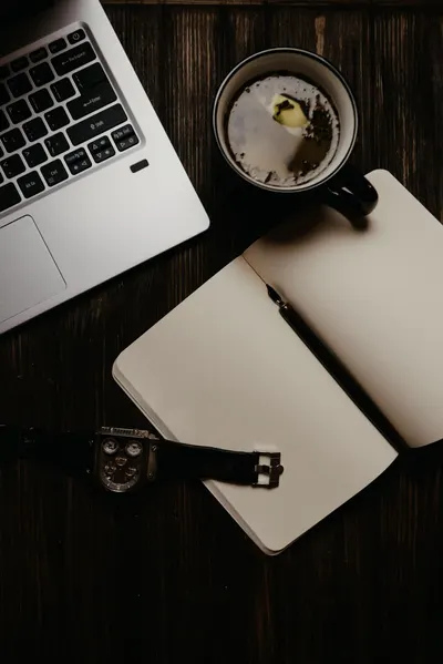 Top view of a laptop, watch, notebook, and cup of tea on a wooden table.