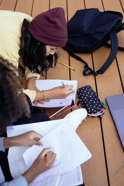 High angle of crop anonymous diverse schoolchildren writing in workbook while doing homework on wooden platform on street