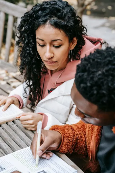 From above of focused ethnic female student sitting at wooden table with black man highlighting text in book while studying together on terrace