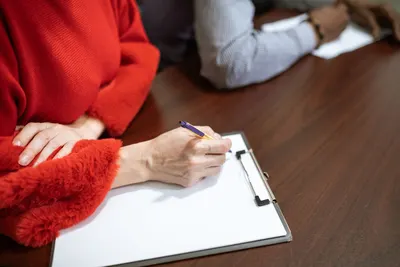 A person in a red sweater writes on a clipboard in a meeting setting.
