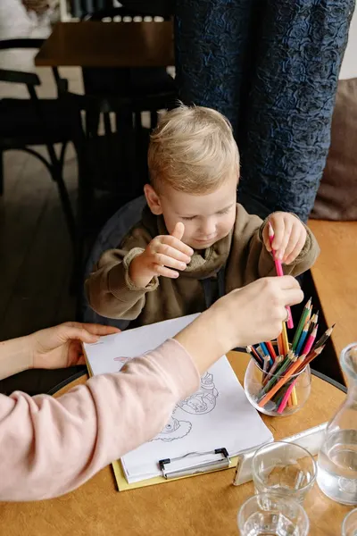 A young boy draws with his parent using colored pencils on a paper indoors.