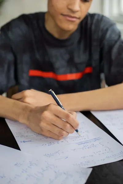 Unrecognizable African American male taking notes on papers and solving equations while sitting at table during studying math lesson in classroom