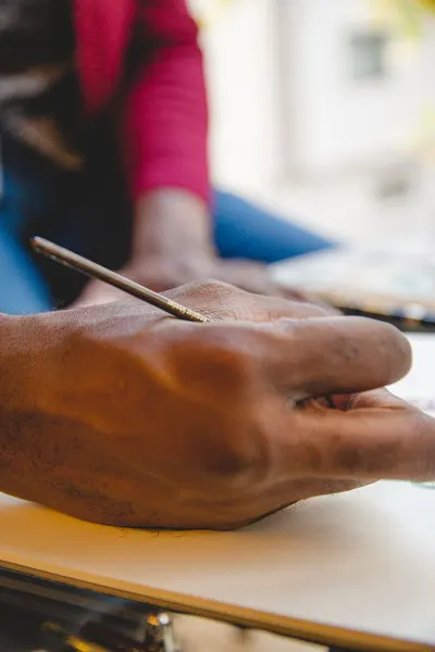Unrecognizable African American male with brush painting while sitting at table on blurred background in light art studio with window