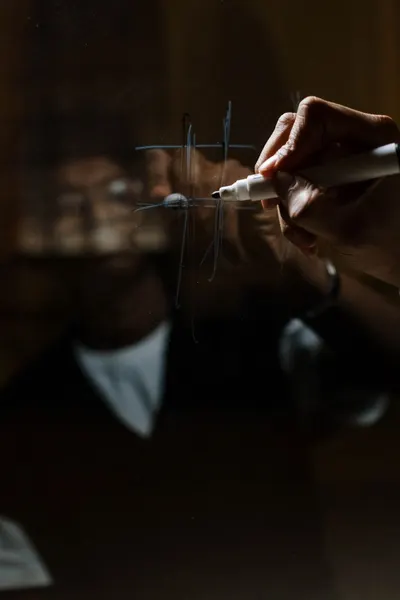 A hand draws a tic tac toe game on glass with a marker, reflection visible.