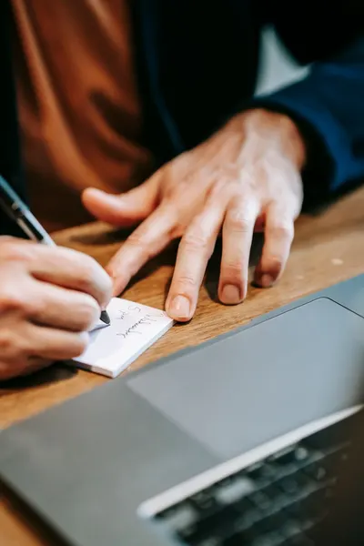 Focused hands writing on paper with laptop on a wooden table; a blend of analog and digital work.