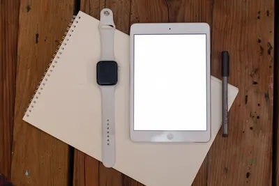 Flat lay of smartwatch, tablet, notepad, and pen on wooden surface.