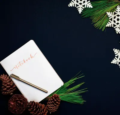 A holiday-themed flat lay featuring a notebook, pinecones, and decorative snowflakes against a dark background.