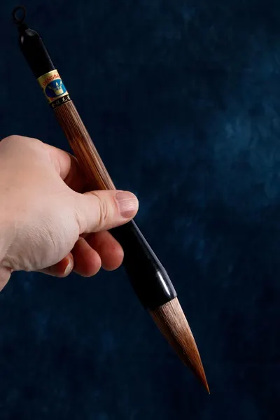 Close-up of a hand holding a traditional calligraphy brush with a blue background.
