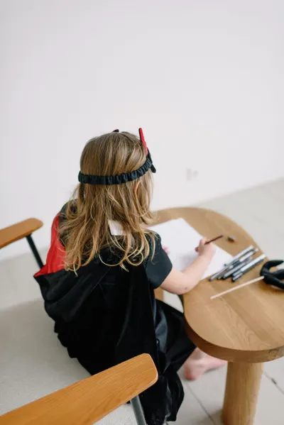 A young girl in a demon costume sits drawing on a wooden table indoors.