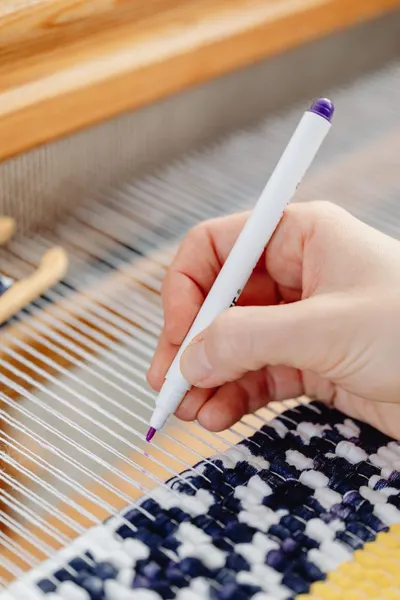 Detailed view of a hand using a marker on a weaving project with colorful yarn.