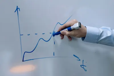 Hand drawing an upward graph on a whiteboard using a blue marker being held by a person in a formal shirt.