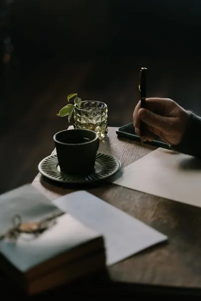 A cozy setting featuring a coffee cup, a hand holding a pen, and a notepad on a wooden table.