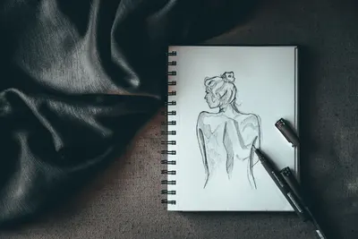 Top view composition of spiral notebook with sketch of undressed female placed on dark surface near liners and black silk fabric