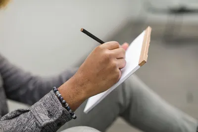 An adult hand writes in a notebook with a pencil, wearing a beaded bracelet and gray plaid sleeves.