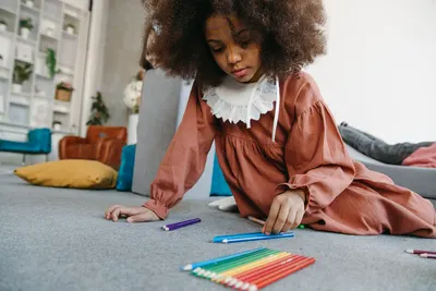 Child wearing a brown dress sitting on the floor with colored pencils indoors.