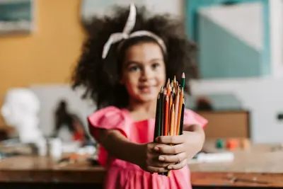 Smiling young girl in pink dress holding colored pencils, showcasing creativity in an art room.