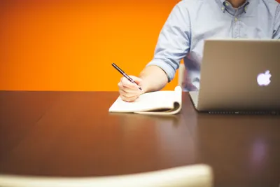 Adult writing notes in a notebook beside a laptop on a wooden desk with orange background.