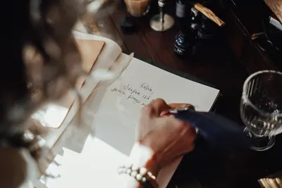 From above of crop anonymous female writer with feather taking notes in notebook at table in sunlight