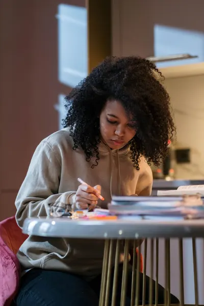 Young woman with afro hair studying at home, focused on textbooks at the kitchen table.